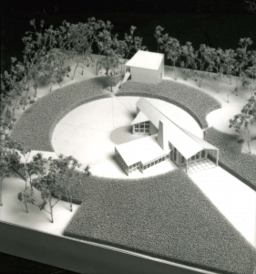 Model of an almost airplane-shaped house with a rectangular extension on one side, with a ring of lawn around it.