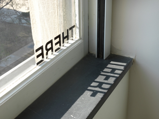The word THERE, in black letters, displayed on a window, its shadow cast on the windowsill