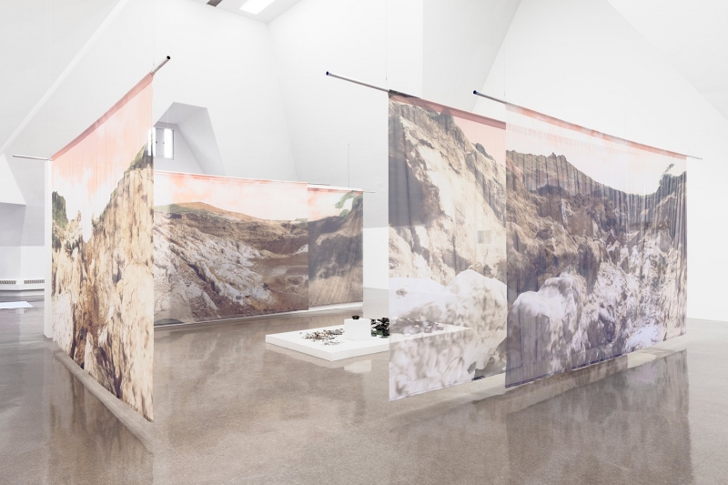 Transparent fabric prints of mountain and desert landscapes with a pink sky around a metal sculpture resting on a platform
