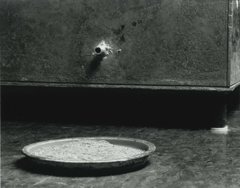 A spigot protrudes from the corner of a large box, opening above a flat dish filled with ashes.