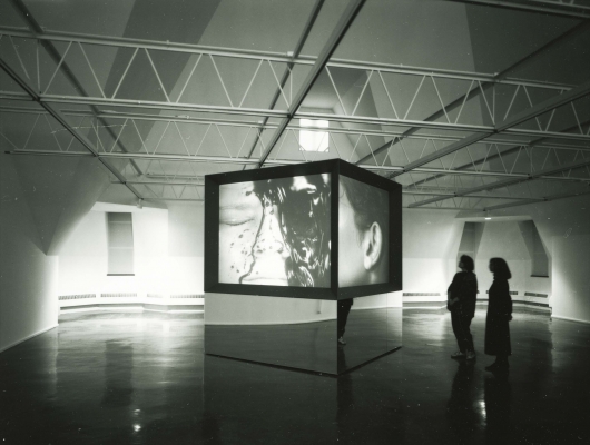 A large cube stands in the gallery, projecting a video of a calm face being covered in a dark liquid.