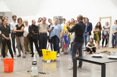A crowd looks on a man gesticulating in front of them, miscellaneous materials strewn around the exhibition space.