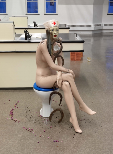A mannequin sits on a toilet seat, wearing a nurse's hat, with a coffee cup and horseshoes dangling from her neck
