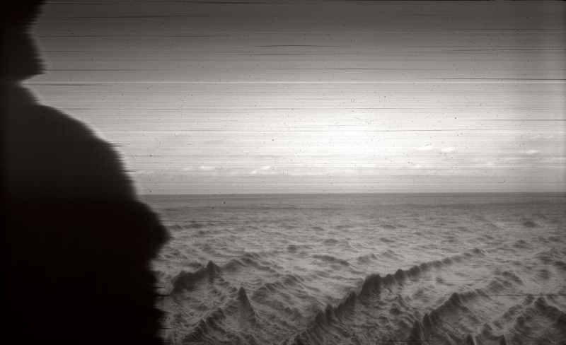A black-and-white photograph of a sparse landscape, looking onto the horizon, its left edge burnt into a curve