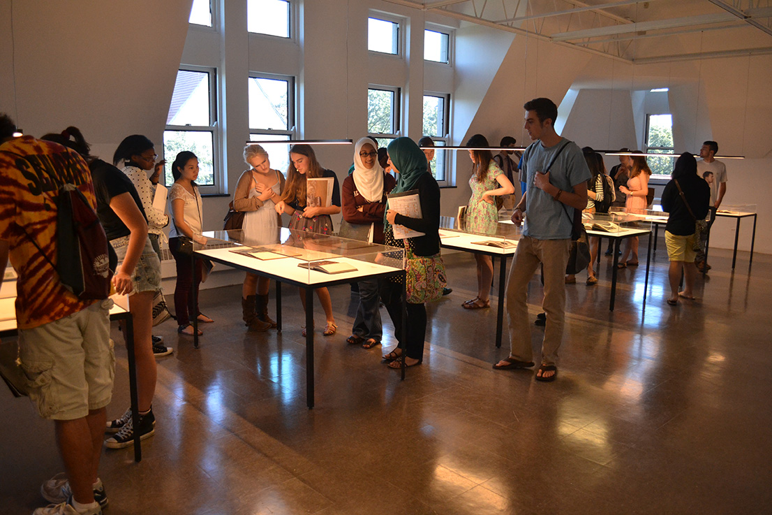 A group of students looks at works displayed on tables under glass.