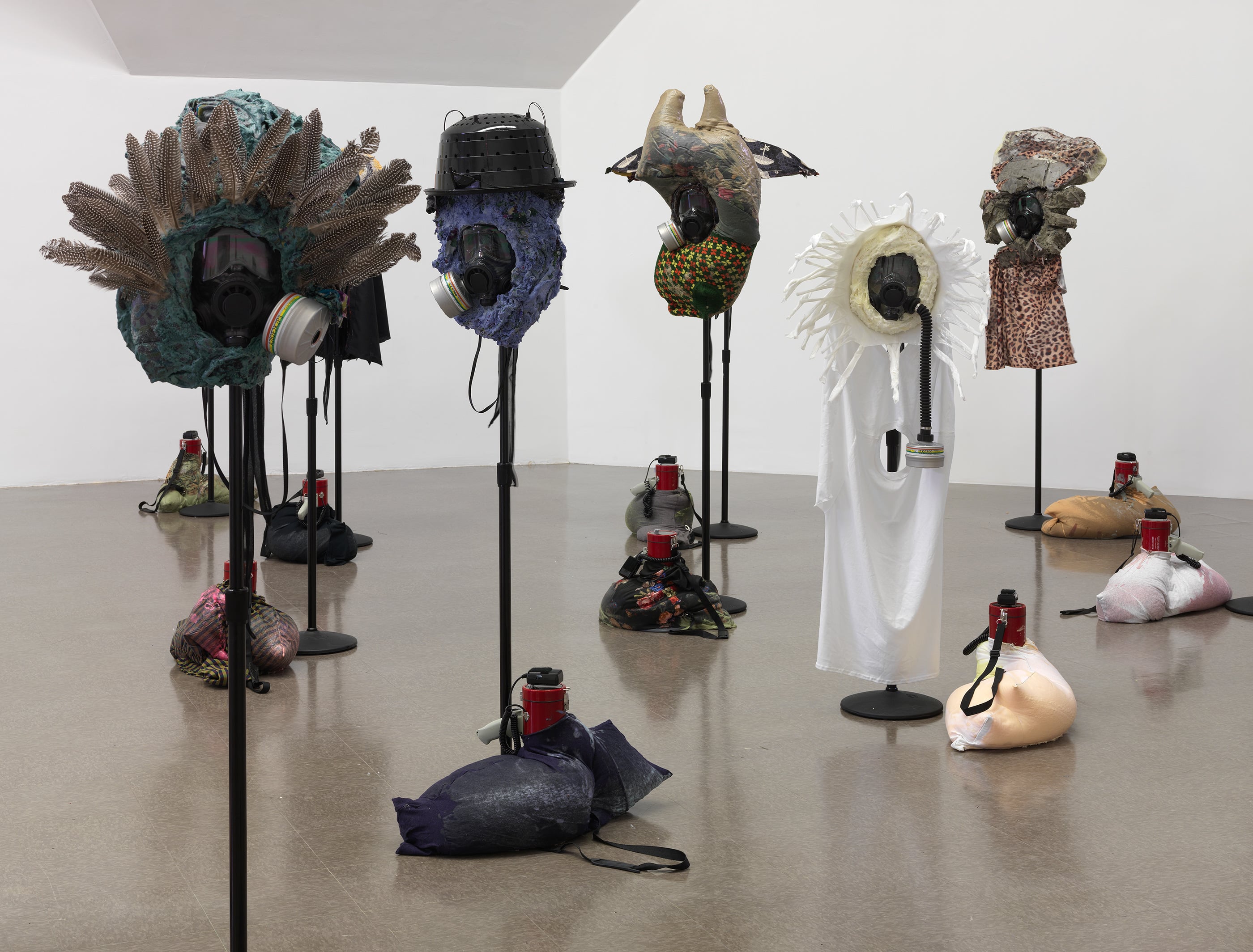 Mixed-media sculptures cover gas masks perched on poles, bags with a megaphone resting at the base of each pole.